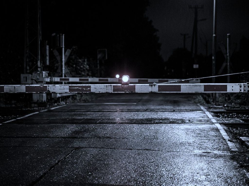 A railway level-crossing barrier lowered across a wet road at night, one red lamp lit.