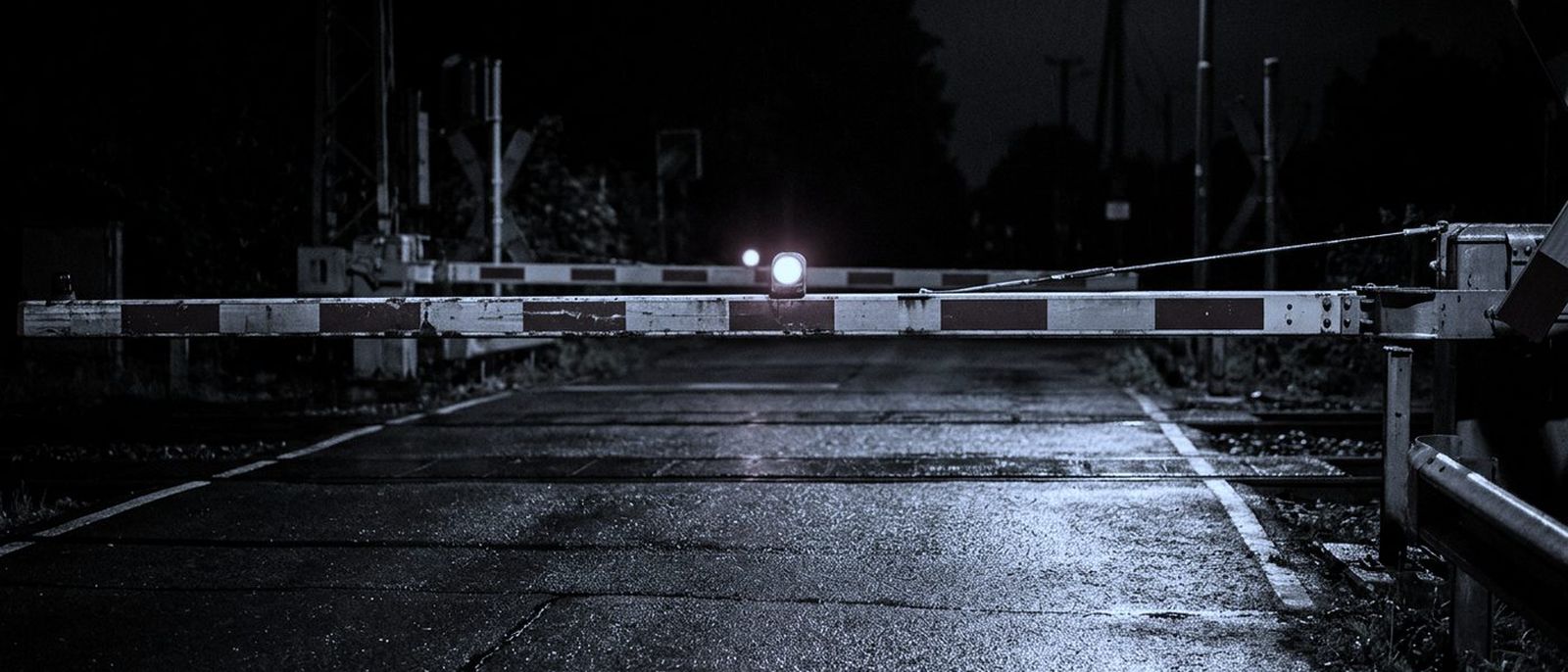 A railway level-crossing barrier lowered across a wet road at night, one red lamp lit.