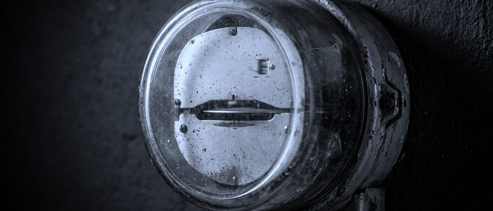 An old analogue electricity meter with its metal disc spinning behind glass on a dark wall.