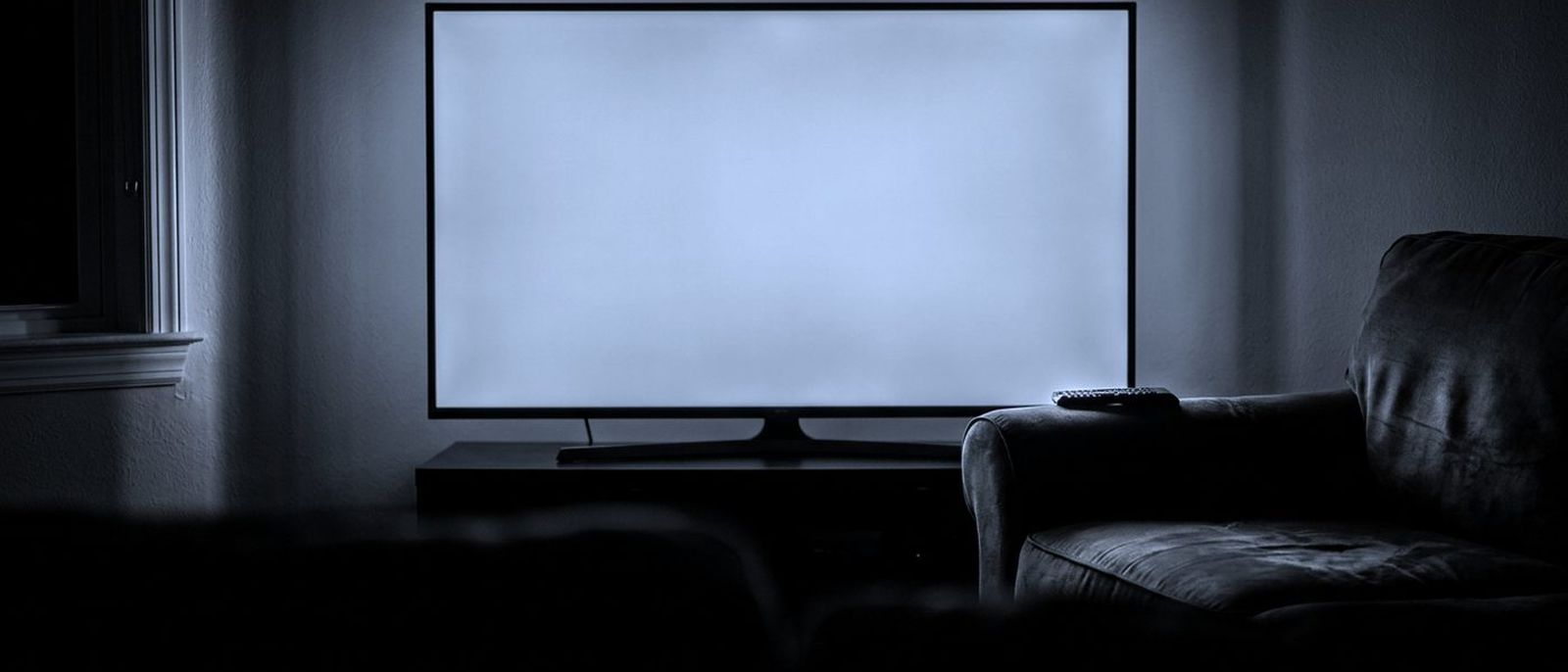 A television glowing in a dark living room, a remote control resting on the sofa arm.