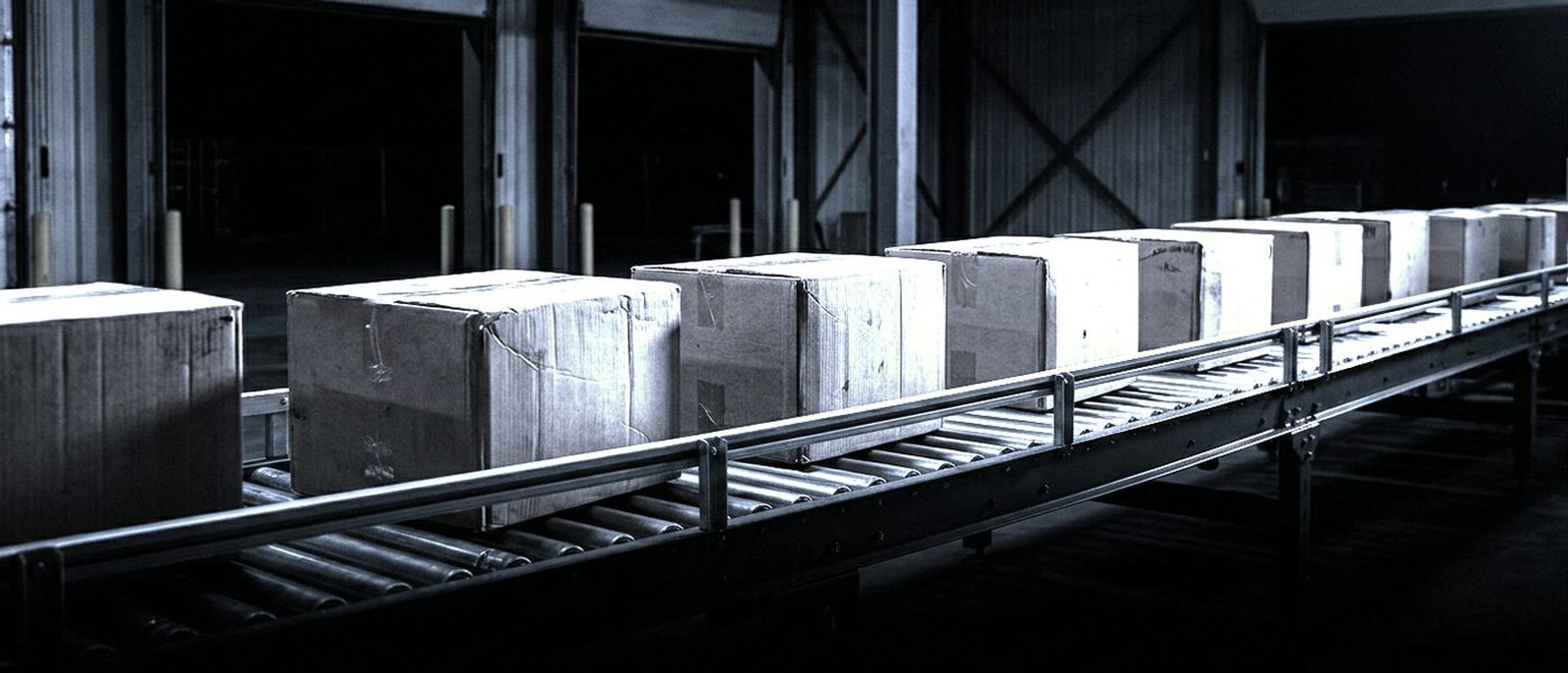 Sealed boxes moving along a conveyor between 2 warehouse bays at night.