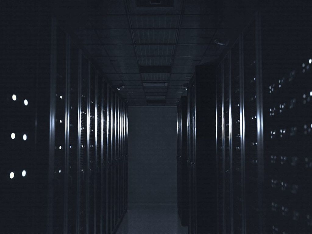 Rows of server racks in a dark data centre aisle, small status lights glowing along each rack.
