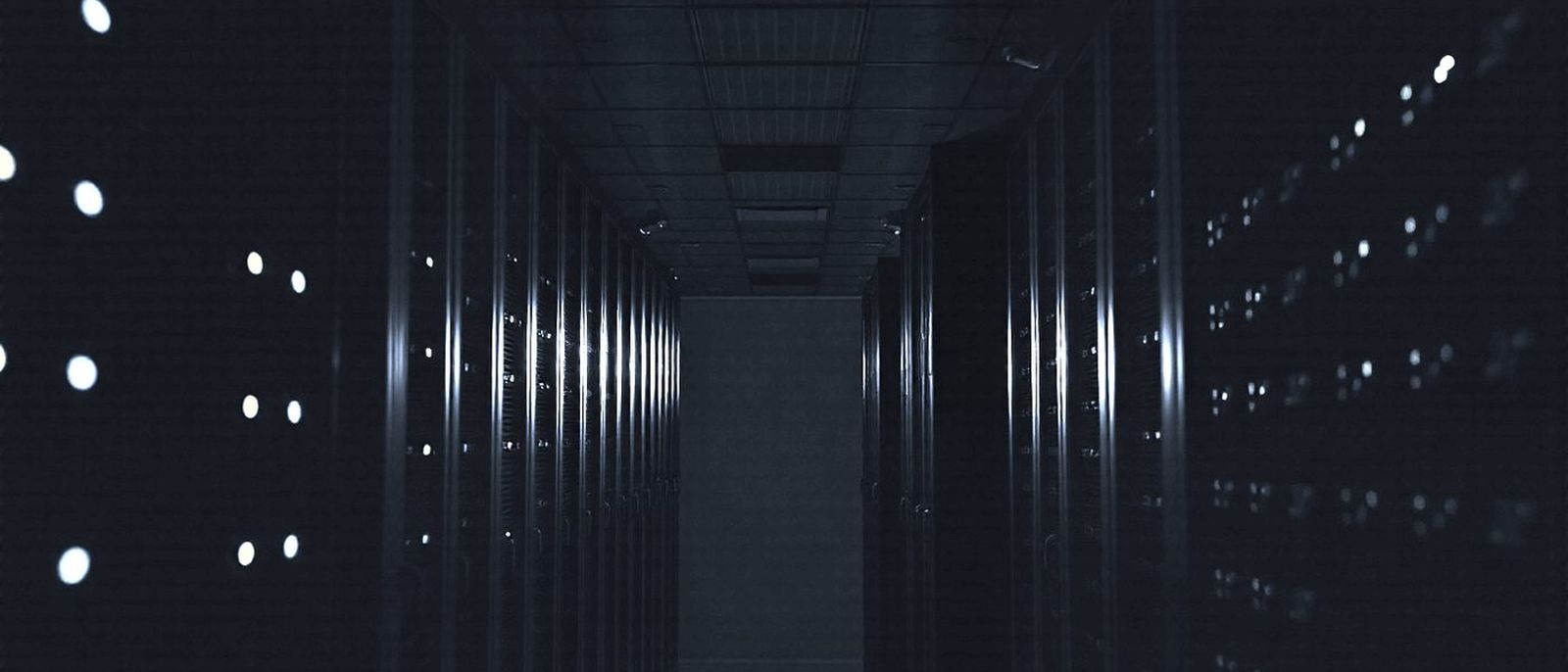 Rows of server racks in a dark data centre aisle, small status lights glowing along each rack.