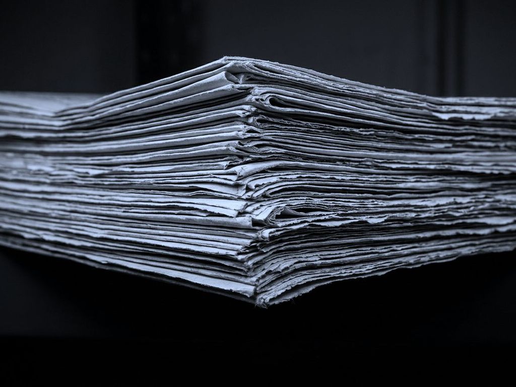 A stack of folded newspapers seen from the side, layered paper edges running the frame.