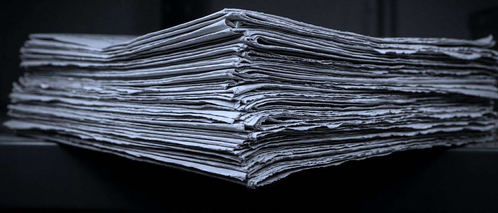 A stack of folded newspapers seen from the side, layered paper edges running the frame.