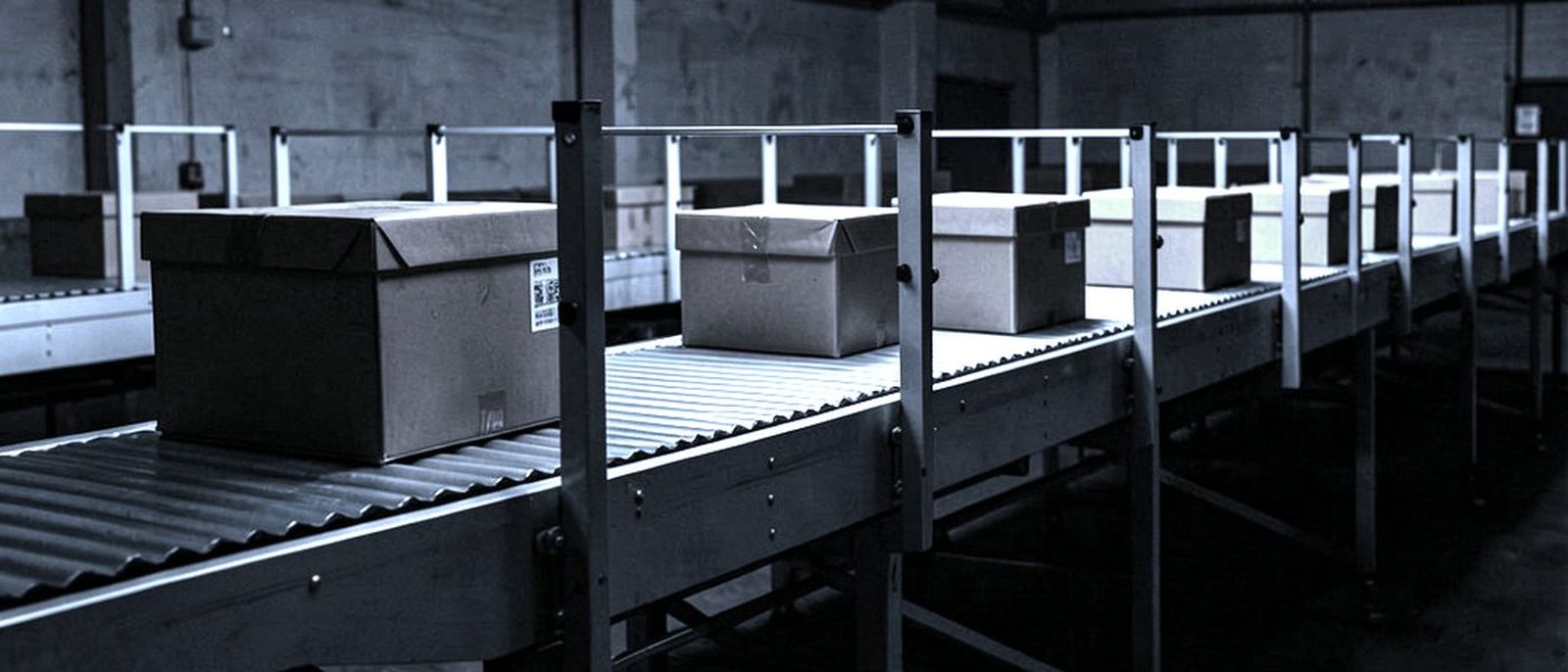 A parcel sorting conveyor in a dark warehouse, boxes moving along the rollers under strip lights.