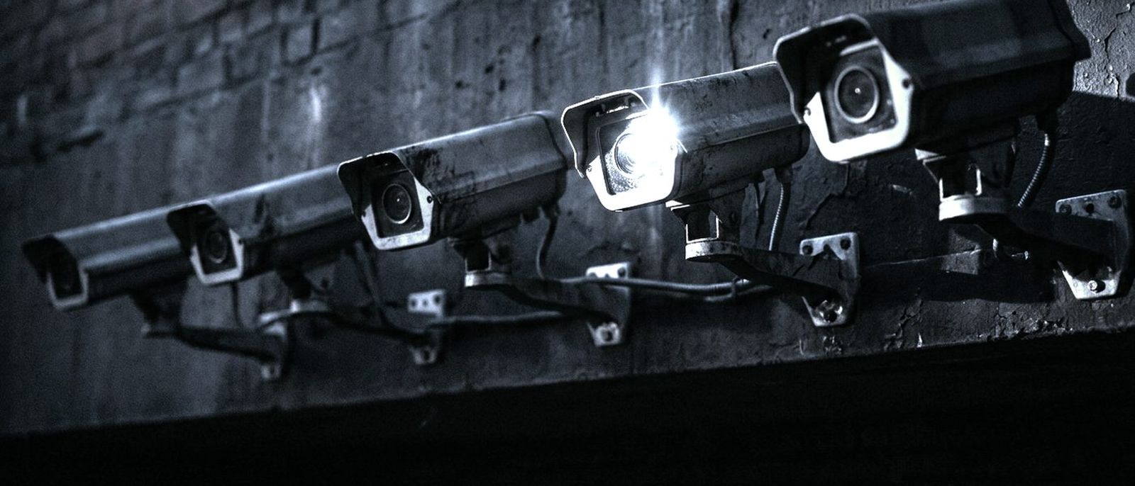 A row of security camera lenses on a dark wall, one catching a reflection of light.