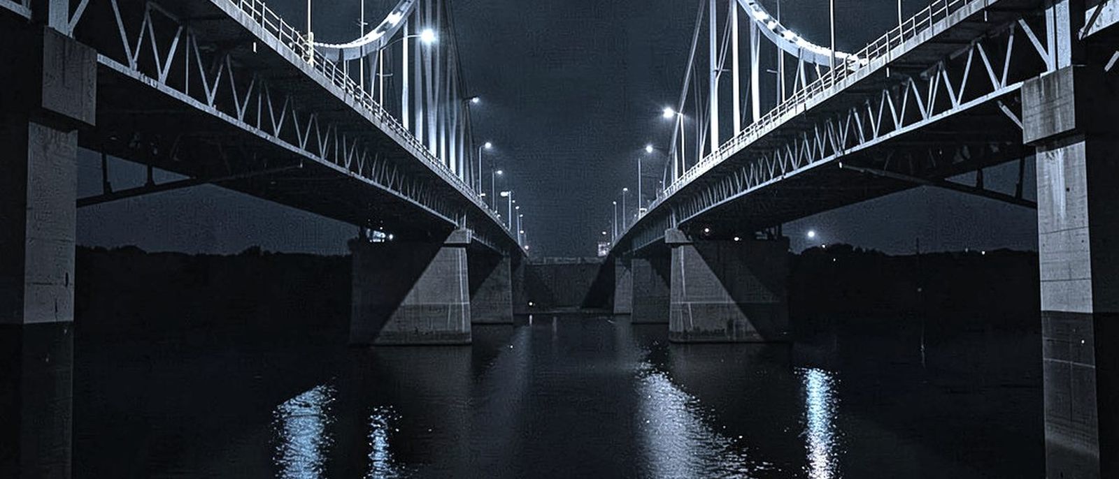 Two bridge spans meeting over dark water at night, lit by lamps along the deck.