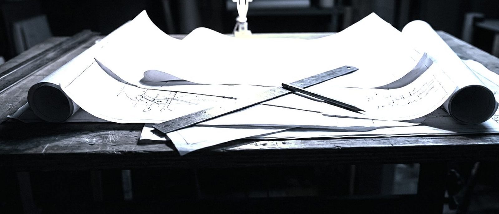 Architectural blueprints unrolled on a drafting table with a steel ruler and a pencil under one lamp.