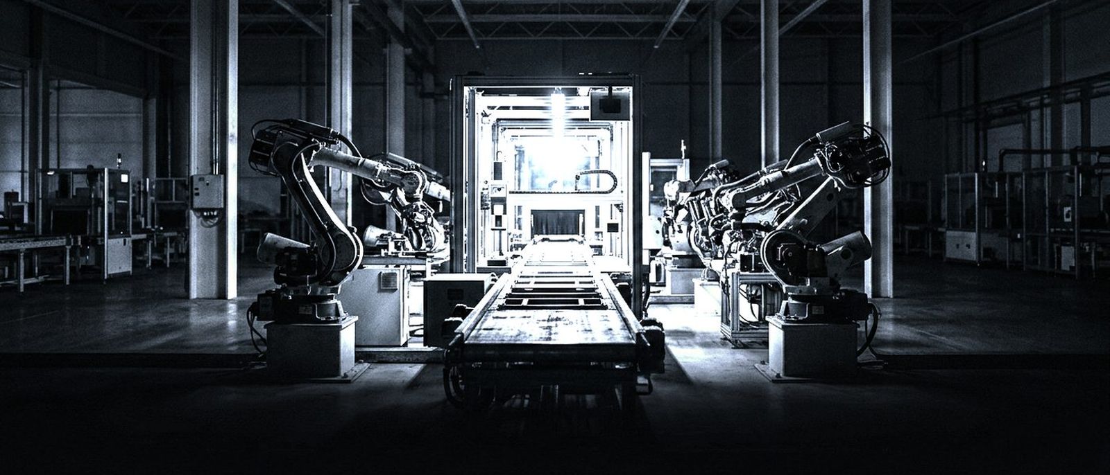 A conveyor line in a dark factory hall, paused at one brightly lit inspection station, robotic arms idle beside it.