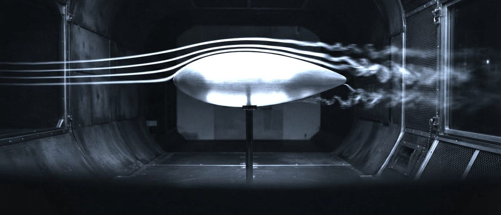 A brushed aluminium test body on a slim mount inside a wind tunnel, smoke lines streaming over it.