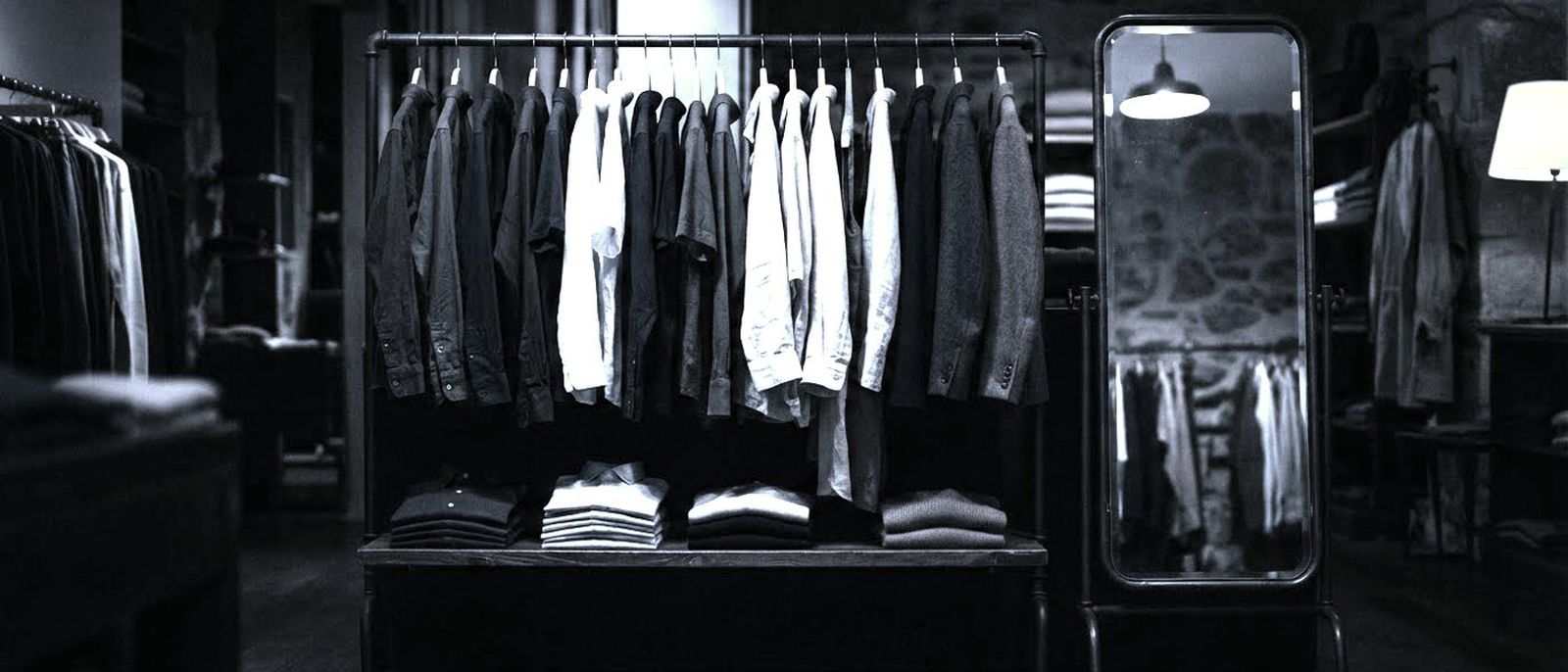A rail of shirts and jackets in a dark boutique, a tall mirror lit softly beside it.