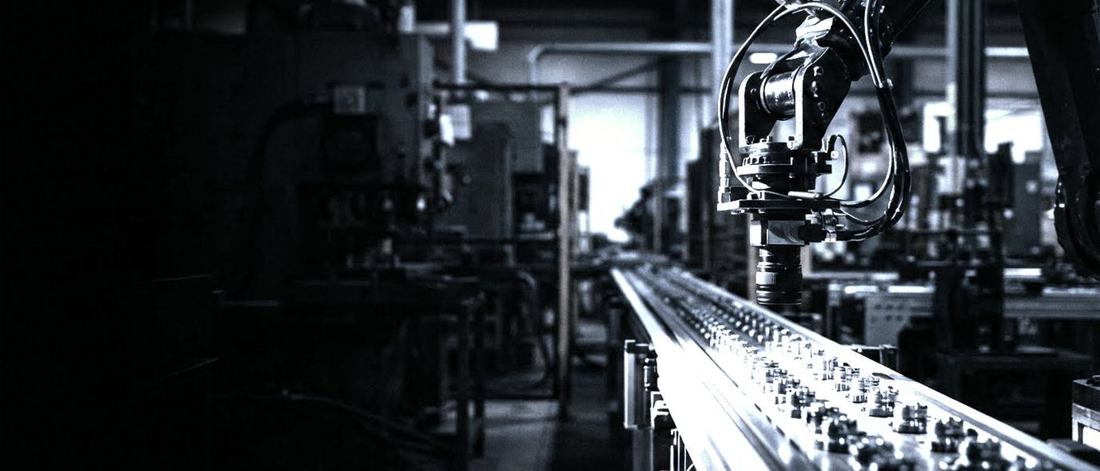 A machine vision camera on an industrial arm above a conveyor of identical parts in a dark factory.