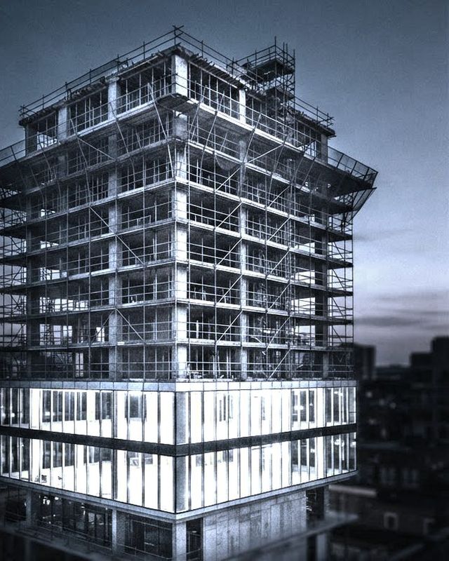A half-finished concrete building wrapped in scaffolding at dusk, the lower floors glazed and lit.